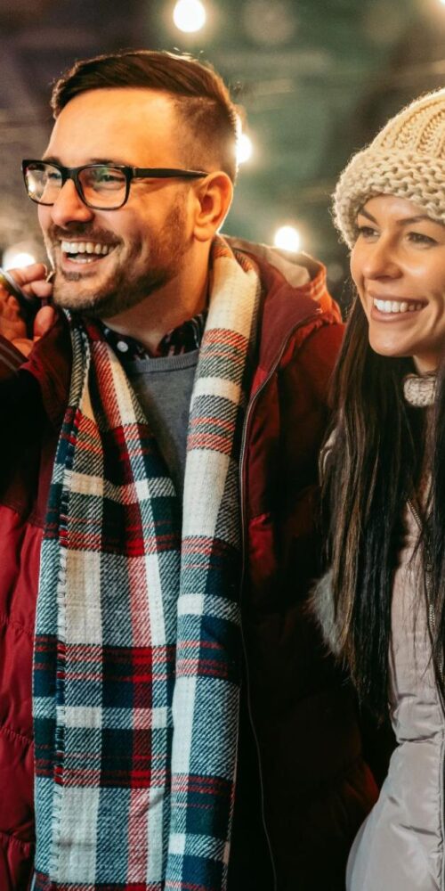 Smiling couple carrying shopping bags at a festive outdoor market during the holiday season.