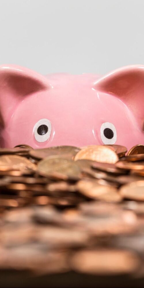 Pink piggy bank partially hidden among a large pile of coins, representing savings or financial goals.