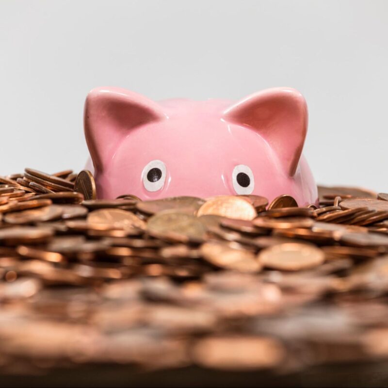 Pink piggy bank partially hidden among a large pile of coins, representing savings or financial goals.