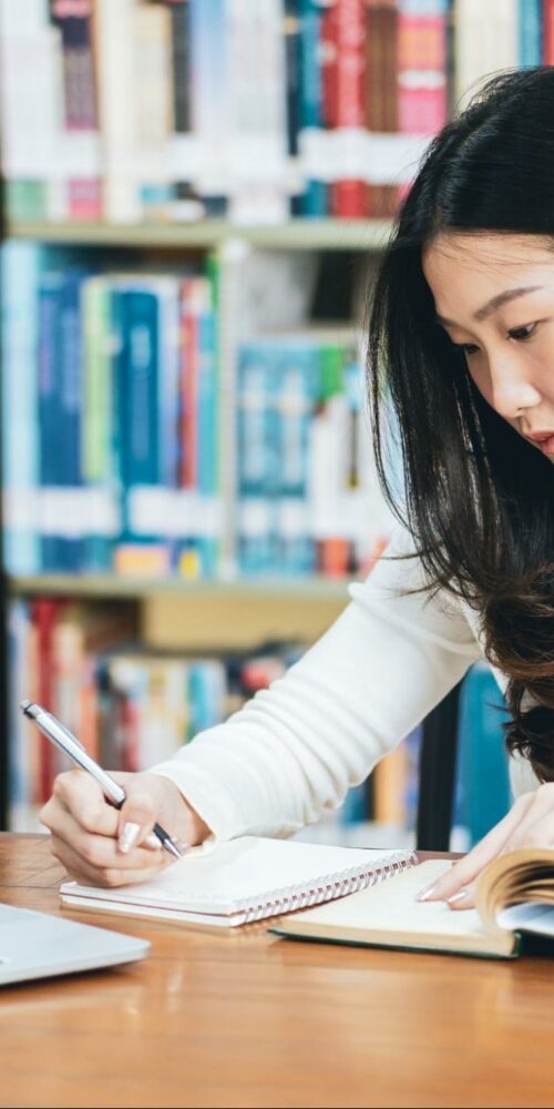 A focused student studying in a library, taking notes from an open book while using a laptop.