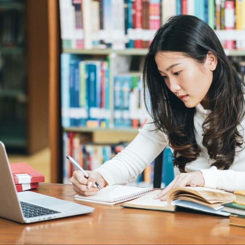 A focused student studying in a library, taking notes from an open book while using a laptop.